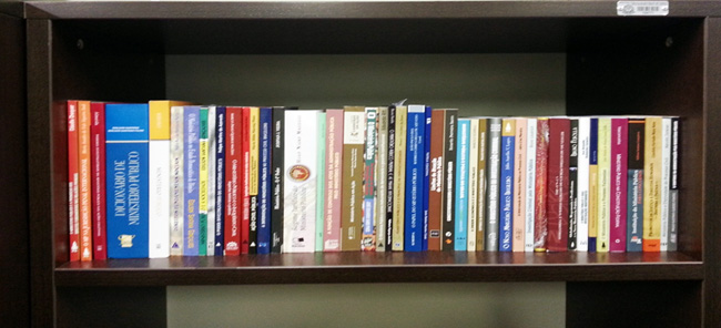 imagem de uma prateleira contendo aproximadamente 30-40 livros, mostrando Parte do acervo de livros do Memorial MPSP sobre o Ministério Público.