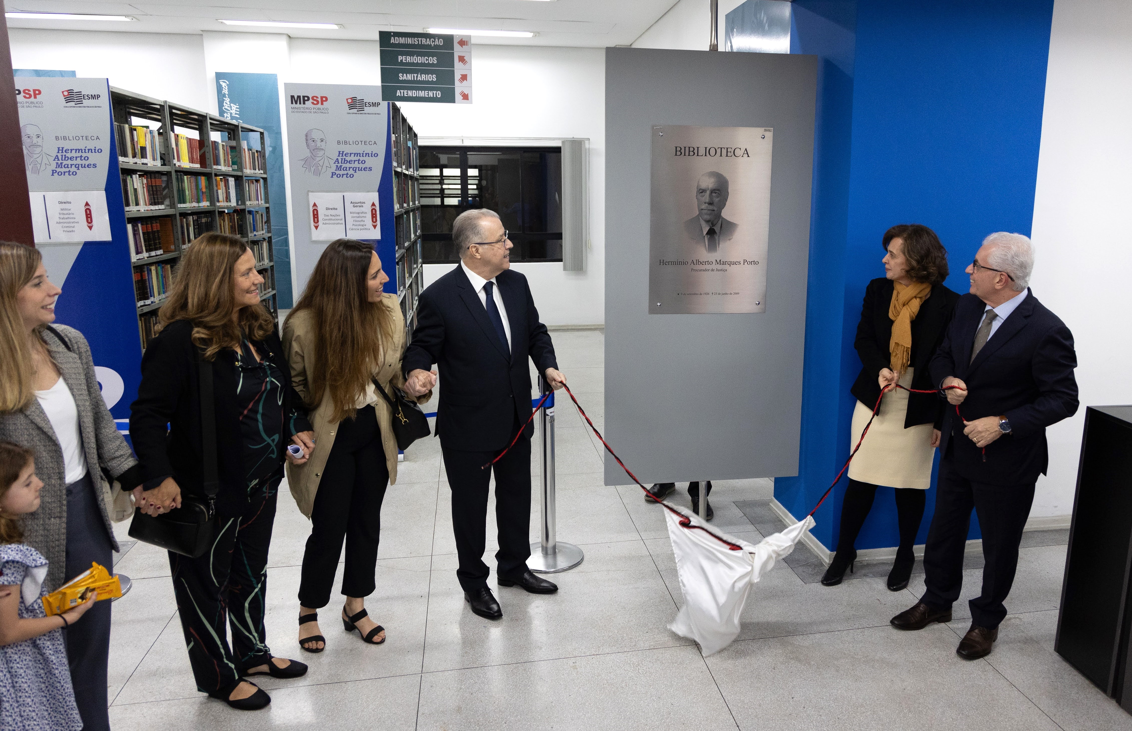 Foto do descerramento de placa, na cerimônia de integração, mostra a diretora da ESMPSP, o PGJ do MPSP e os filhos e netas do homenageado. Eles estão de pé em frente à placa da biblioteca. Os familiares estão de mãos dadas. O PGJ e a diretora da ESMPSP seguram a corda do tecido que cobria a placa. Fim da descrição. 